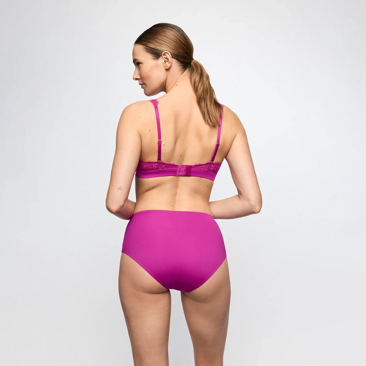 A woman shown from the back wearing a bright magenta two-piece lingerie set, consisting of a lace bralette and high-waisted briefs, against a light gray studio background.