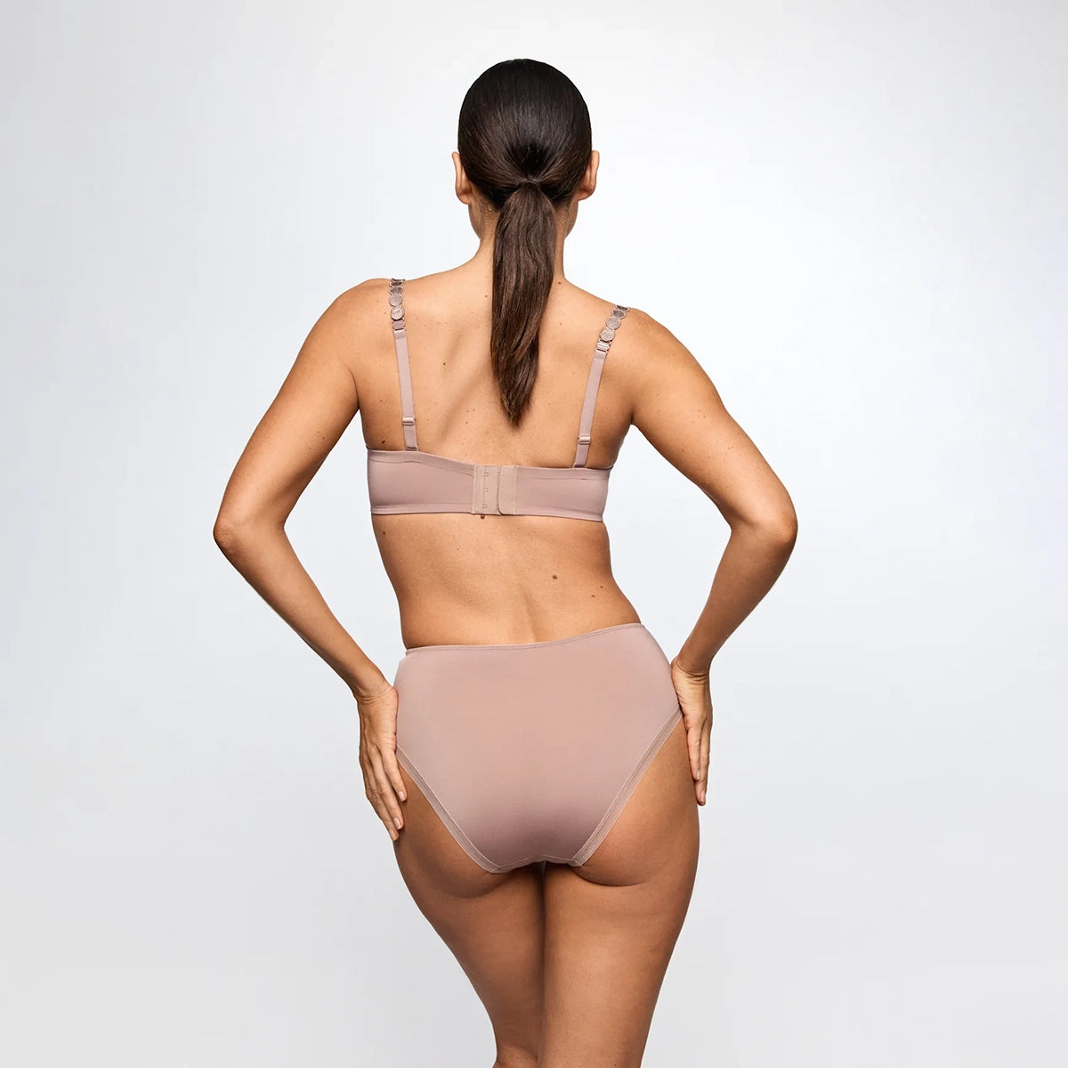 A woman shown from the back wearing a matching nude-beige bra and high-waisted brief set against a light gray studio background.