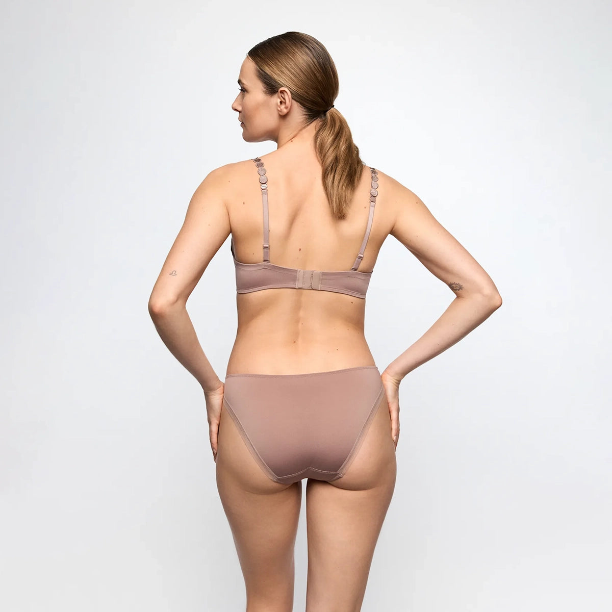 A woman shown from the back wearing a matching two-piece underwear set in a muted taupe-brown color, consisting of a bralette and high-waisted briefs, against a plain light gray studio background.