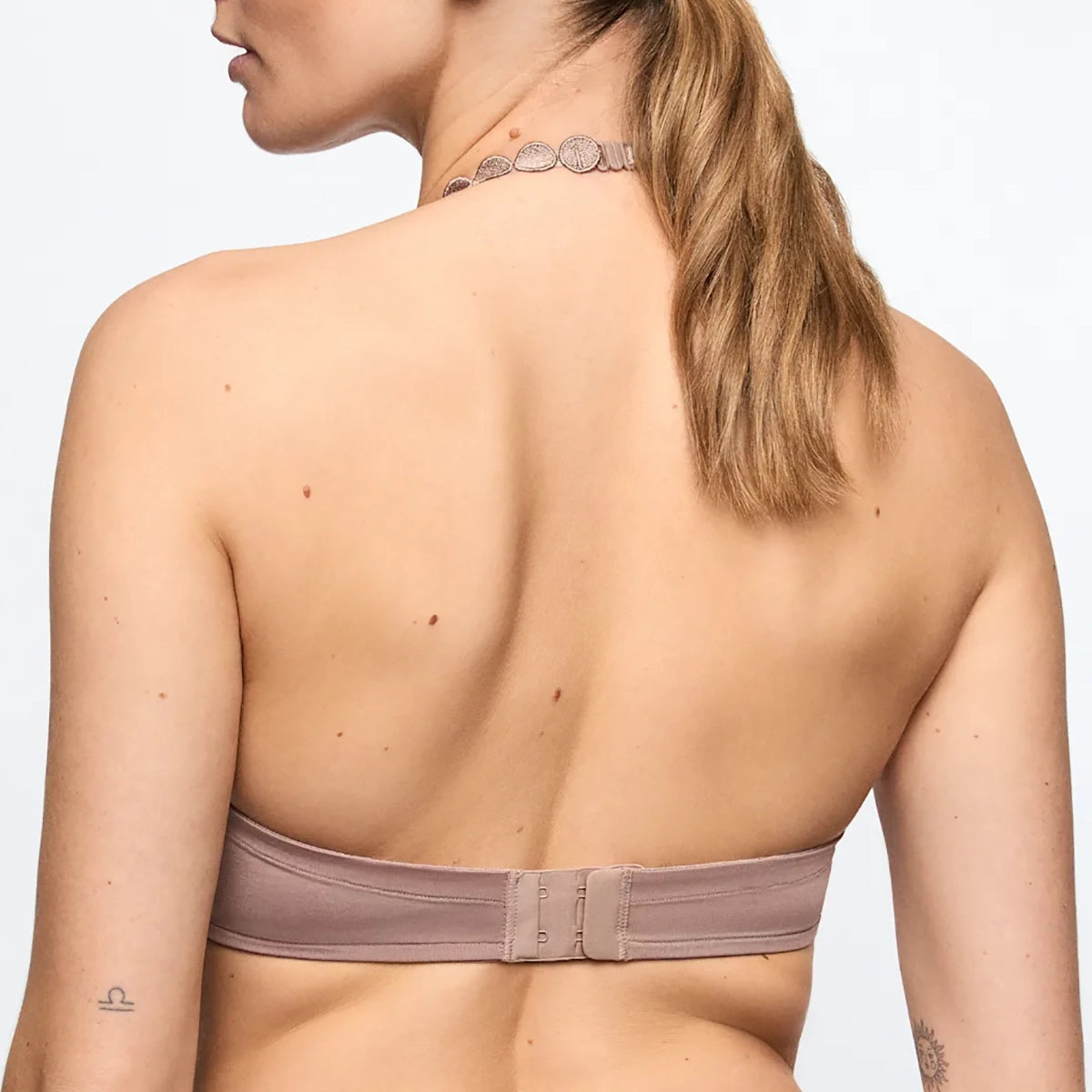 Rear view of a woman wearing a strapless bandeau-style bra in a muted mauve-taupe color, with a matching thin chain necklace visible against a plain light background.