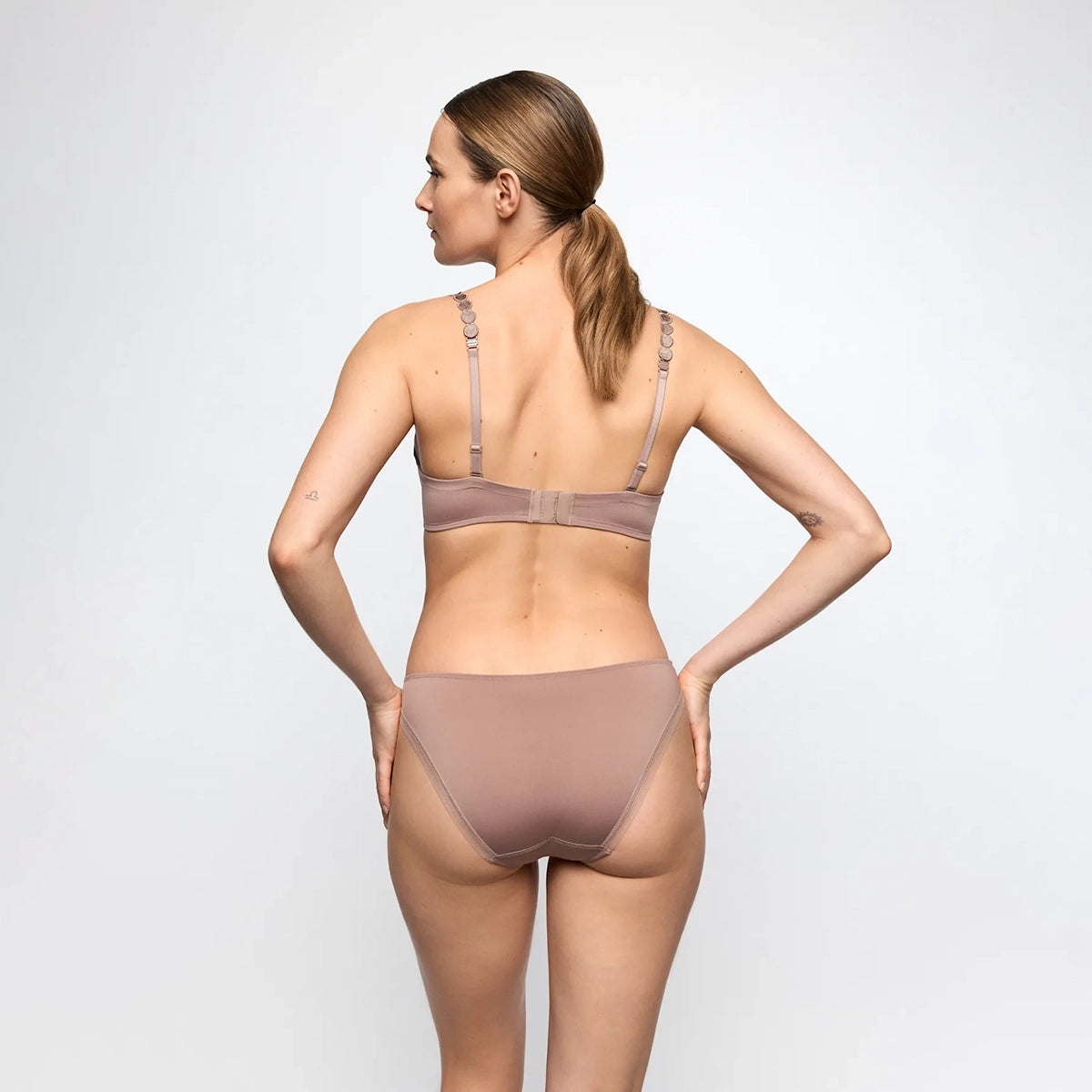 A woman shown from the back wearing a matching two-piece underwear set in a muted taupe-brown color, consisting of a bralette and high-waisted briefs, against a plain light gray studio background.