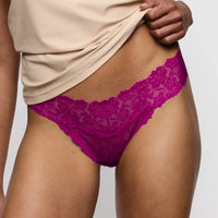 A close-up of a model wearing bright magenta lace thong underwear, with a beige top being lifted to reveal the underwear. The background is a clean, neutral light gray.