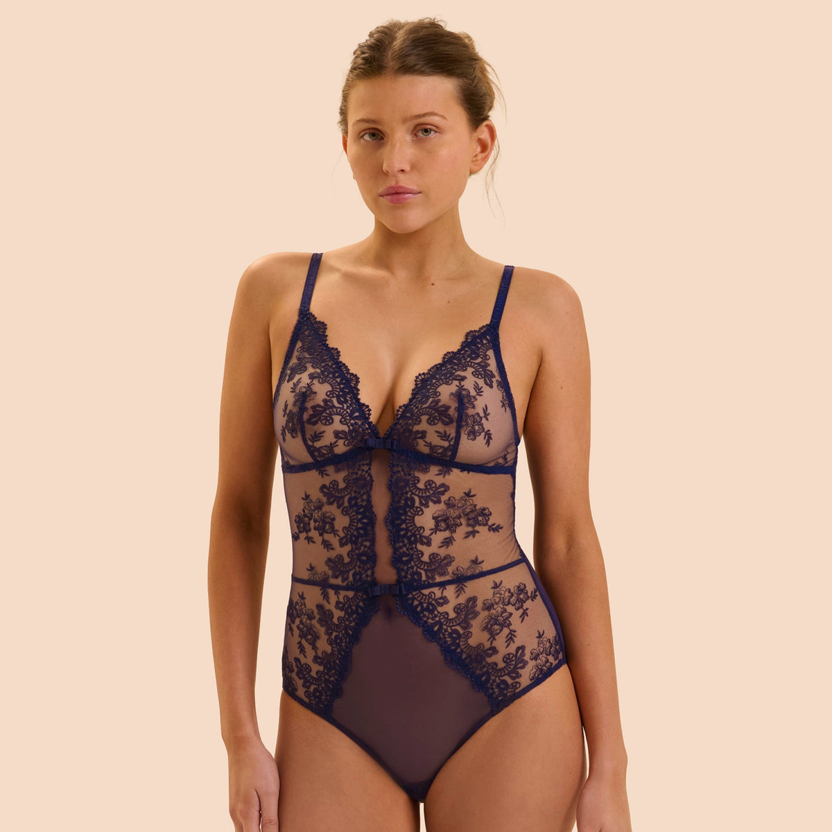 A woman modeling a dark navy blue sheer lace bodysuit with floral lace patterns and solid mesh panels, set against a solid beige background.