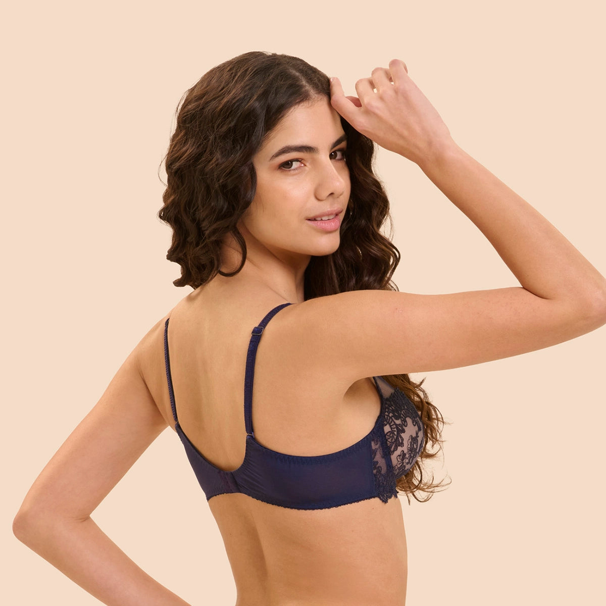 A woman modeling a navy blue lace-trimmed underwire bra, shown from the back against a solid light beige background.