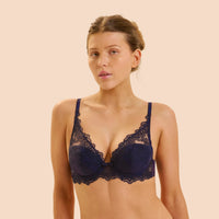 A woman modeling a dark navy blue lace bra against a solid pale peach background.