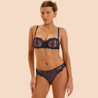 A woman modeling a dark navy blue lace lingerie set against a solid pale peach background. The set includes a sheer underwire bra with floral lace cups and matching lace-trimmed briefs.