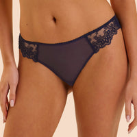 A close-up of a model wearing dark navy blue sheer mesh and lace panties against a solid light beige background.